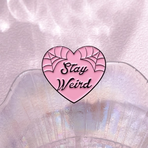 Pink heart enamel pin with Stay Weird text and spider web details on metal base