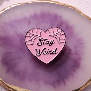 Pink heart enamel pin with Stay Weird text and spider web details on agate coaster