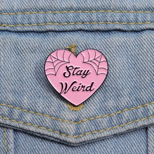 Close-up of pink heart enamel pin with Stay Weird text on a blue denim jacket pocket