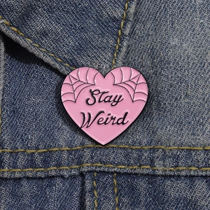 Pink heart enamel pin with Stay Weird slogan and spiderweb on denim jacket