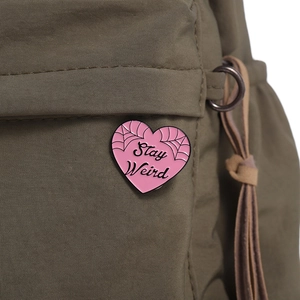 Detail of pink heart enamel pin with Stay Weird text on olive green fabric bag