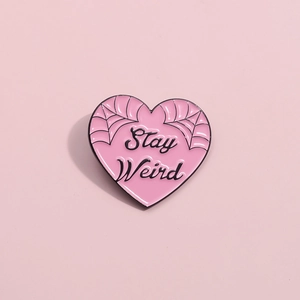 Pink heart enamel pin with Stay Weird text and spider web patterns on a clean background