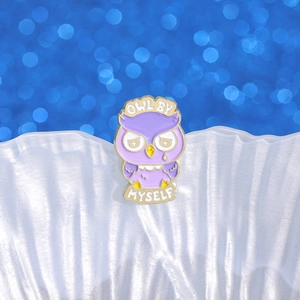 Purple owl enamel brooch with 'OWL BY MYSELF' slogan on a sparkling blue bokeh background