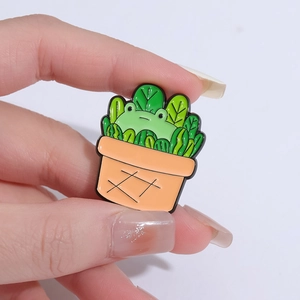 Handheld view of a green frog in an orange flower pot enamel pin with vibrant leaves