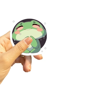 Hand holding a cute green cartoon frog sticker for guitar and laptop decoration