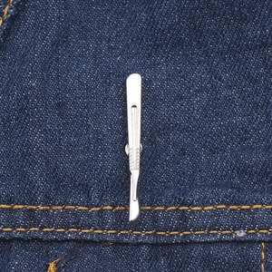 Dettaglio spilla a forma di bisturi argento con impugnatura su tessuto denim blu