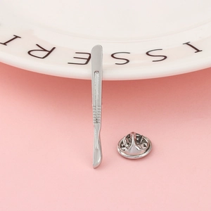 Spilla in metallo a forma di bisturi argento lucido per medici su sfondo rosa