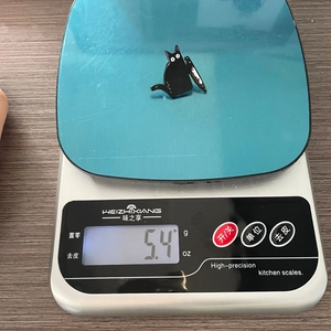 Detailed shot showing a 5.4g cartoon black killer cat enamel pin on a digital scale