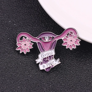 Enamel uterus brooch with flowers, eye design, and 'Grow a Pair' text on black texture