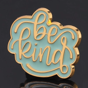Macro shot of gold-plated enamel pin with light blue finish and 'be kind' artistic script