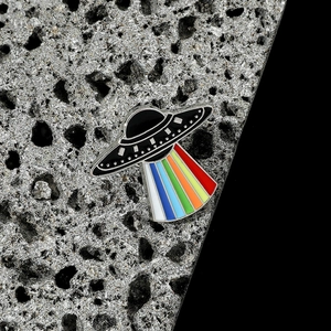 Detailed view of UFO enamel pin with vibrant rainbow light beam on grey rock surface