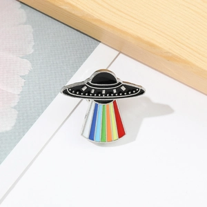 Close-up of a UFO enamel pin featuring a colorful rainbow beam on a white surface