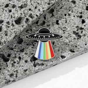 Detail view of a UFO enamel pin with a colorful rainbow beam on a rock background