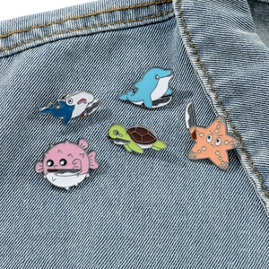 Detail of five funny cartoon ocean animal enamel pins with knives on blue denim fabric