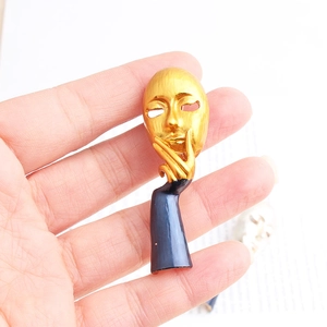 Artistic gold face mask brooch with blue enamel handle held by hand over text background