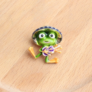 Detailed close-up of a green frog enamel pin with a dotted hat and purple flower on wood