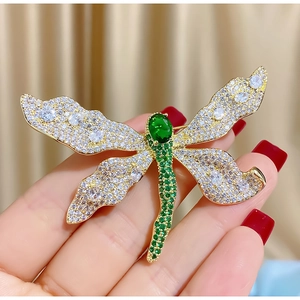 Elegante spilla a libellula dorata con pietre verdi e cristalli su uno sfondo beige