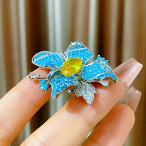 Broche fleur argentée avec pétales bleus et gemme jaune tenue entre les doigts