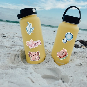 Yellow vacuum water bottles on sand decorated with cute pink axolotl cartoon stickers
