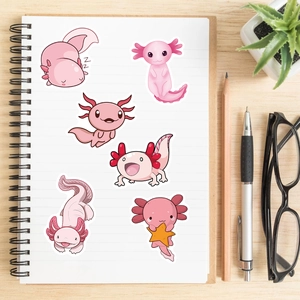 Pink cartoon axolotl stickers displayed on a white notebook with lifestyle props