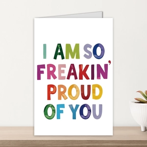 Tarjeta de felicitación con texto I AM SO FREAKIN PROUD OF YOU en estante de madera