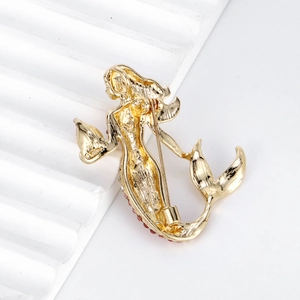 Golden mermaid brooch with red crystals shown from a 45-degree angle on a white stand
