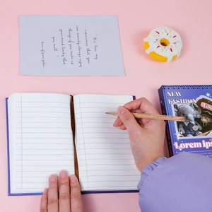 Close-up of hand writing in a navy blue journal with a pencil on a pink aesthetic desk