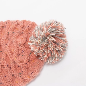 Detail einer Zopfstrickmütze mit dreifarbigem Bommel in Grau, Weiß und Rosa oben