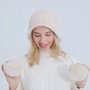 Dettaglio di maglia a trecce e grandi pompon su un cappello invernale color crema con paraorecchi