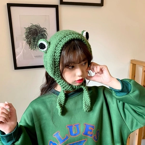 Close-up of green knitted frog earflap hat with large 3D eyes and braided straps