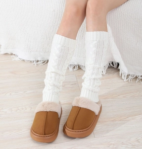 Detail of white cable knit leg warmers paired with brown plush slippers on wood floor