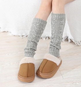Close-up of gray cable-knit leg warmers with brown faux fur lined slippers on wood floor