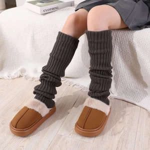 Détail de jambières en tricot gris portées avec des chaussons en peluche marron sur tapis