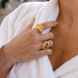 Detalle de anillo dorado con textura de lava y anillo de corazón hueco en modelo con bata
