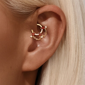 Dettaglio macro del piercing Daith dorato con pietra marquise rossa e catenine pendenti