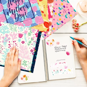 Flat lay of hands decorating Mother's Day scrapbook with floral pattern papers on white wood