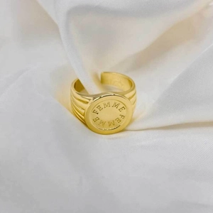 Detalle de un anillo de sello dorado con grabado FEMME y rayas sobre seda blanca