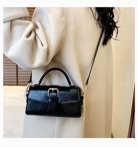 Detail of a black glossy pleated leather handbag with a gold buckle on a beige coat