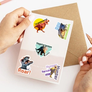 Close-up of hand holding colorful dinosaur stickers with Roar and Hunter text for scrapbooking