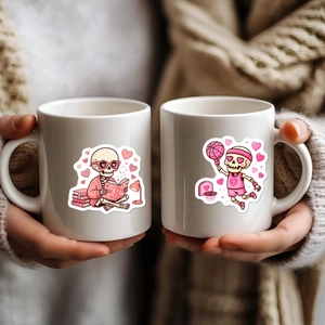 Close-up of ceramic mugs featuring pink cartoon skeleton and heart stickers on a cozy background