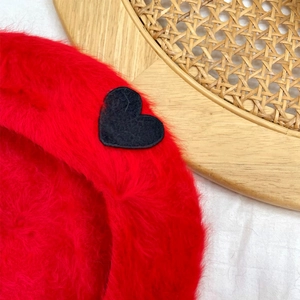 Close-up of red plush beret featuring a soft furry texture and black heart patch detail