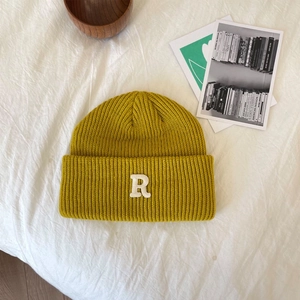 Detail van een mosterdgele ribgebreide muts met een witte letter R patch op een witte stofgrond