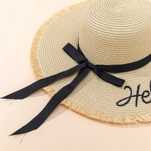 Detail of straw hat with Hello embroidery, black bow ribbon, and fringe brim finish