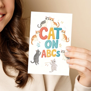 Close-up of a hand holding a cute cat illustration greeting card with CAT ON ABCS text