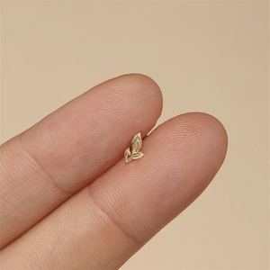 Macro detail of tiny gold leaf cartilage stud earrings held between fingertips for scale