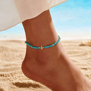 Detail of turquoise beaded anklet with golden cross charm on a sandy beach setting