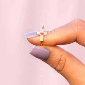 Pink heart and enamel cross ring in gold finish held by hand on purple backdrop