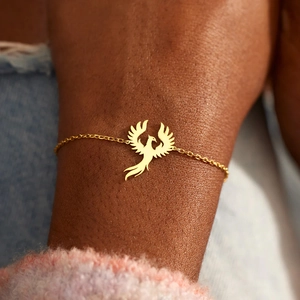 Detailaufnahme eines goldenen Phönix-Charm-Armbands an einem Model mit kuscheligem Strickpullover