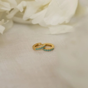 Gold huggie earrings with turquoise row stones on a soft white floral background