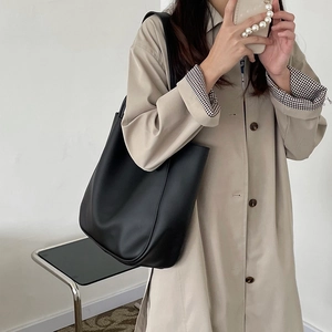 Minimalist large black tote bag in matte faux leather worn by a model in a beige trench coat